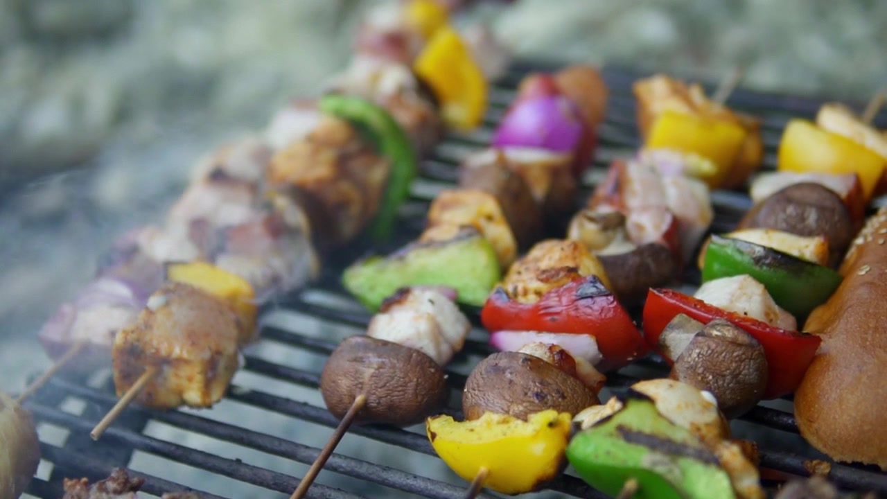 Primo piano di hamburger sfrigolanti e spiedini di verdure vivaci su una griglia esterna, catturando l'atmosfera del barbecue estivo con fiamme fumose e dettagli appetitosi.