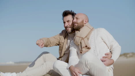 Medium-Shot-Of-A-Cheerful-Gay-Couple-Hugging-And-Talking-Together-While-Sitting-On-The-Beach-And-Looking-Into-Distance