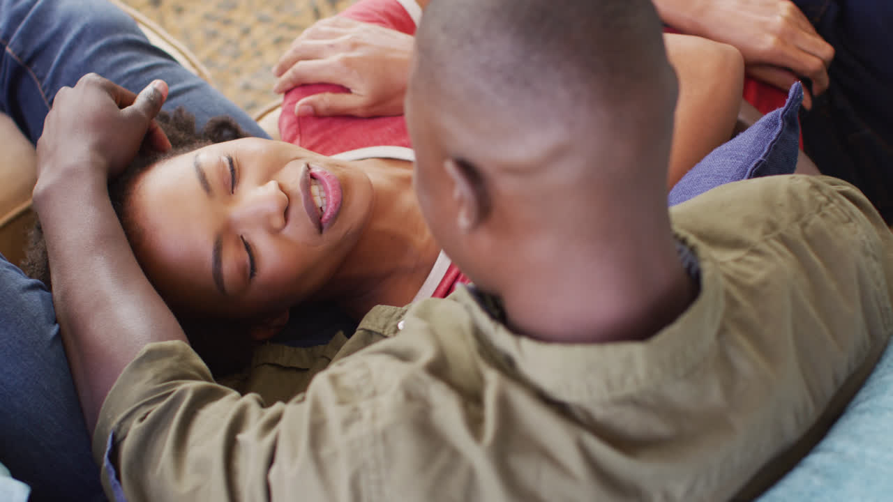 Video of happy african american couple sitting on sofa, talking and embracing