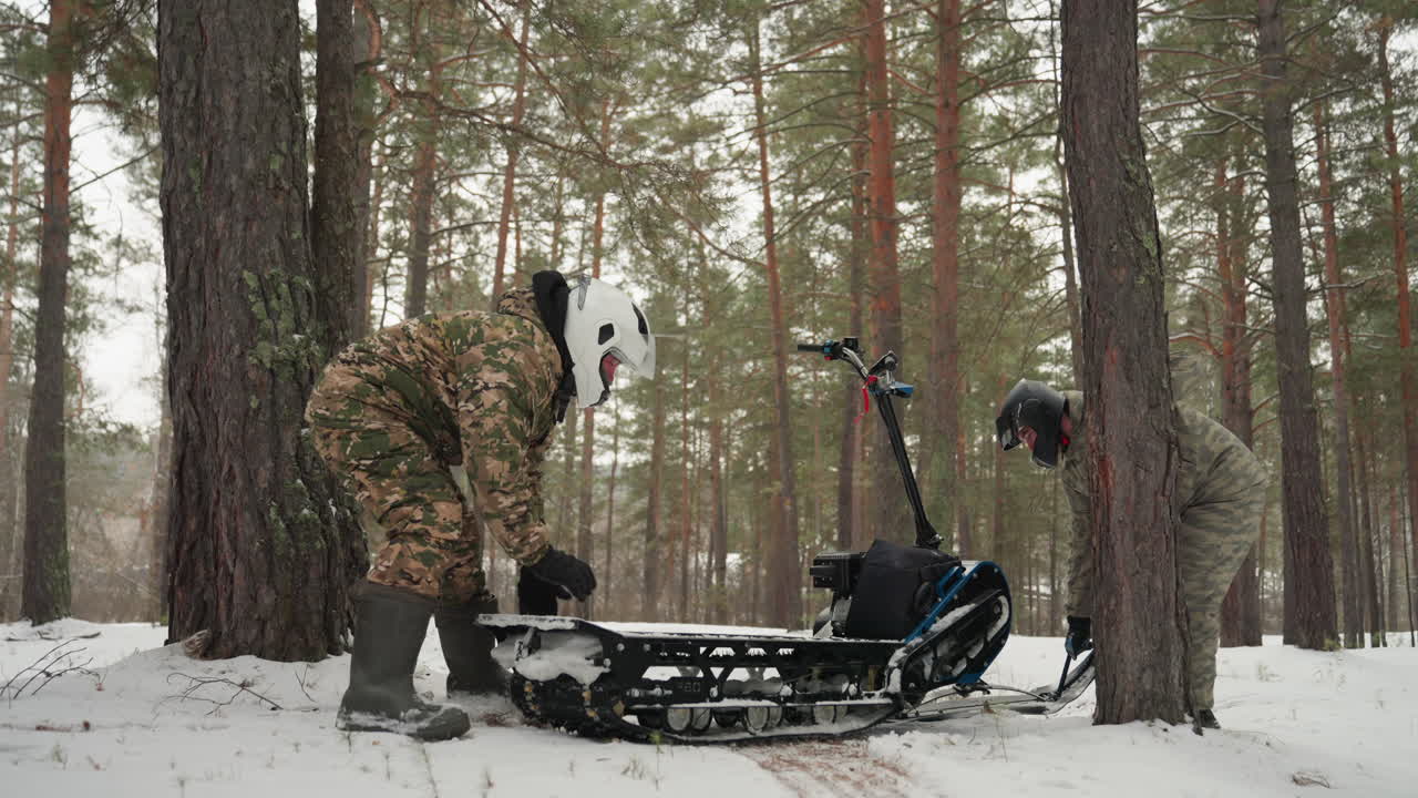 Piloti in abbigliamento bianco riparano l'ATV nella neve, giacche mimetiche caschi bianchi, foresta di pini ghiacciata, motoslitta cingolata incastrata contro un albero, sforzo manuale e lavoro di squadra, respiro visibile e luce invernale smorzata, urgente.