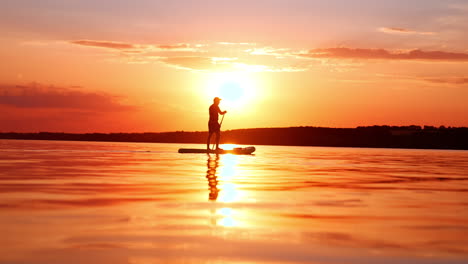 lonely man going sup boarding on the river at sunset. orange sky and waterscape with black silhouette of a man rowing on sup board.