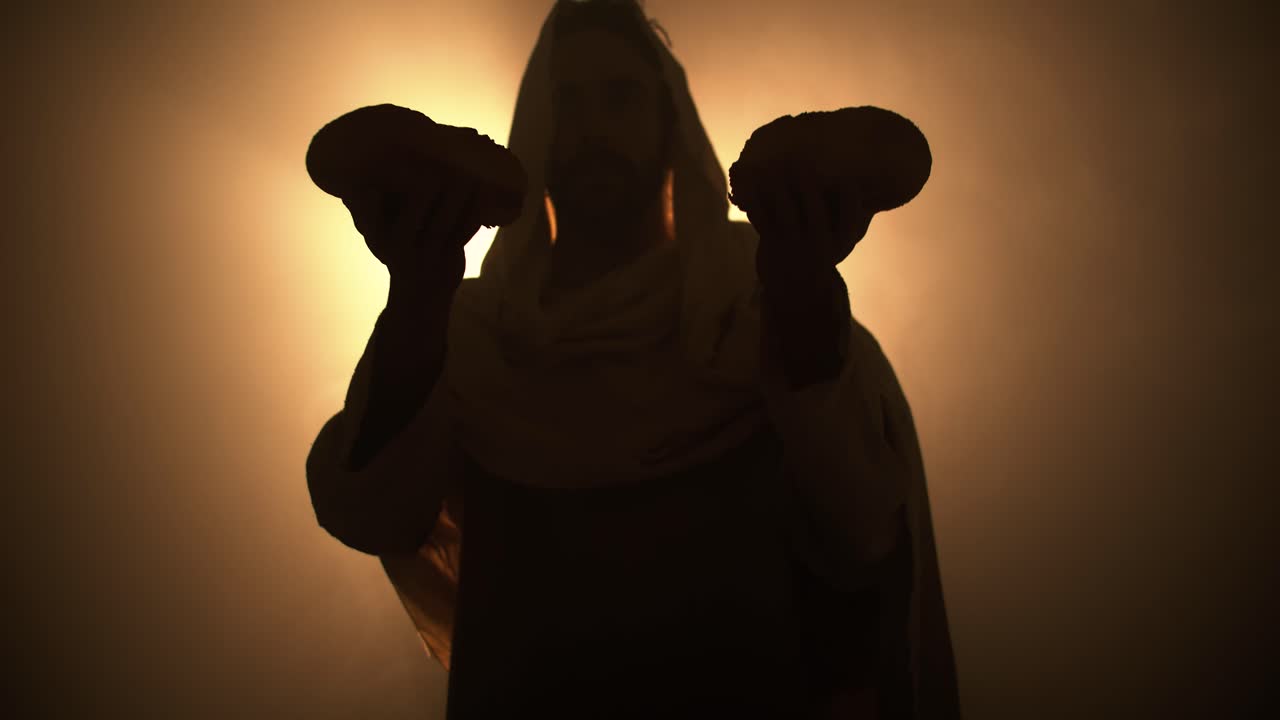 Silhouette of Jesus Christ in white hooded robe praying with bread as he stands in glowing, bright light praying. Representation of Jesus at last supper, communion, Christian Easter resurrection.