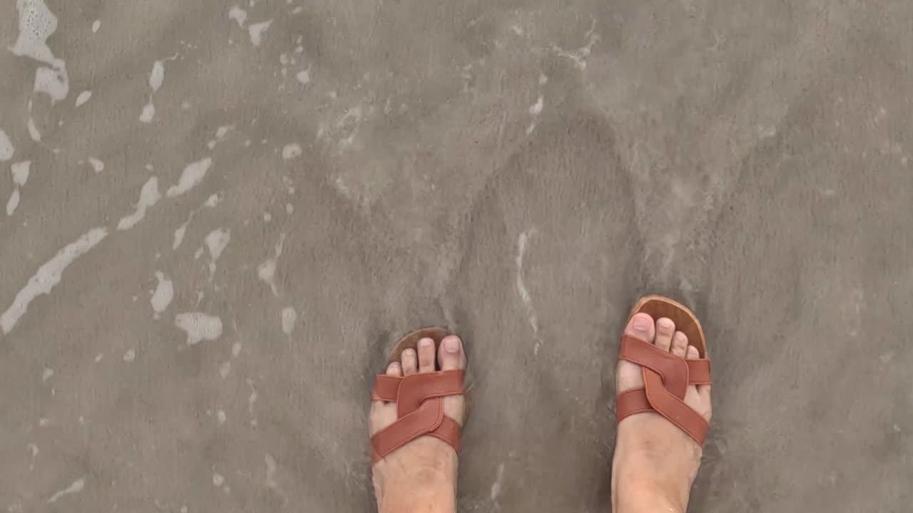 Una prospettiva in prima persona di piedi che indossano sandali marroni, mentre la schiuma del mare lambisce la sabbia