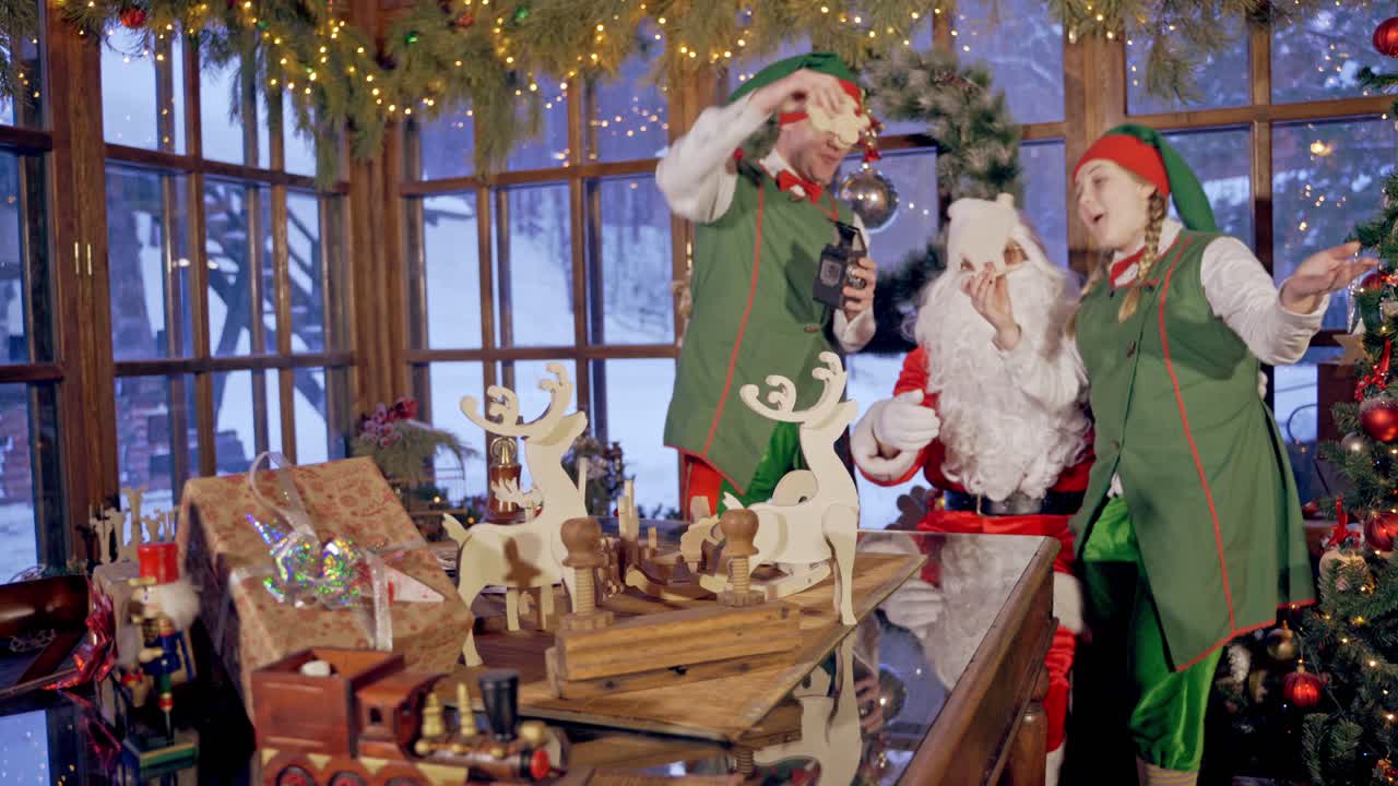 Happy elves with wooden toys. Many handmade toys on the table. Santa Claus having joyful time with fairy characters in the Christmas decorated room.
