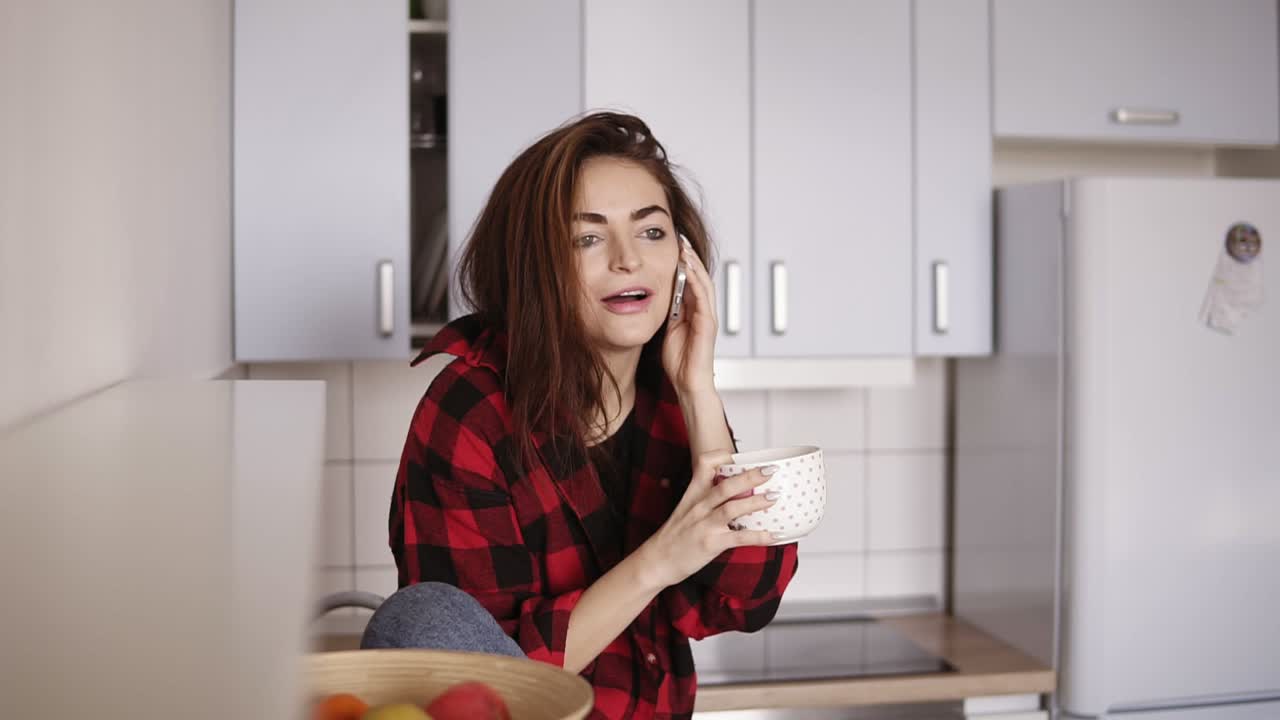 Young beautiful girl starts calling someone while sitting in her kitchen and having a cup of tea in her hand.