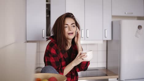 una joven hermosa comienza a llamar a alguien mientras está sentada en su cocina y tiene una taza de té en la mano.
