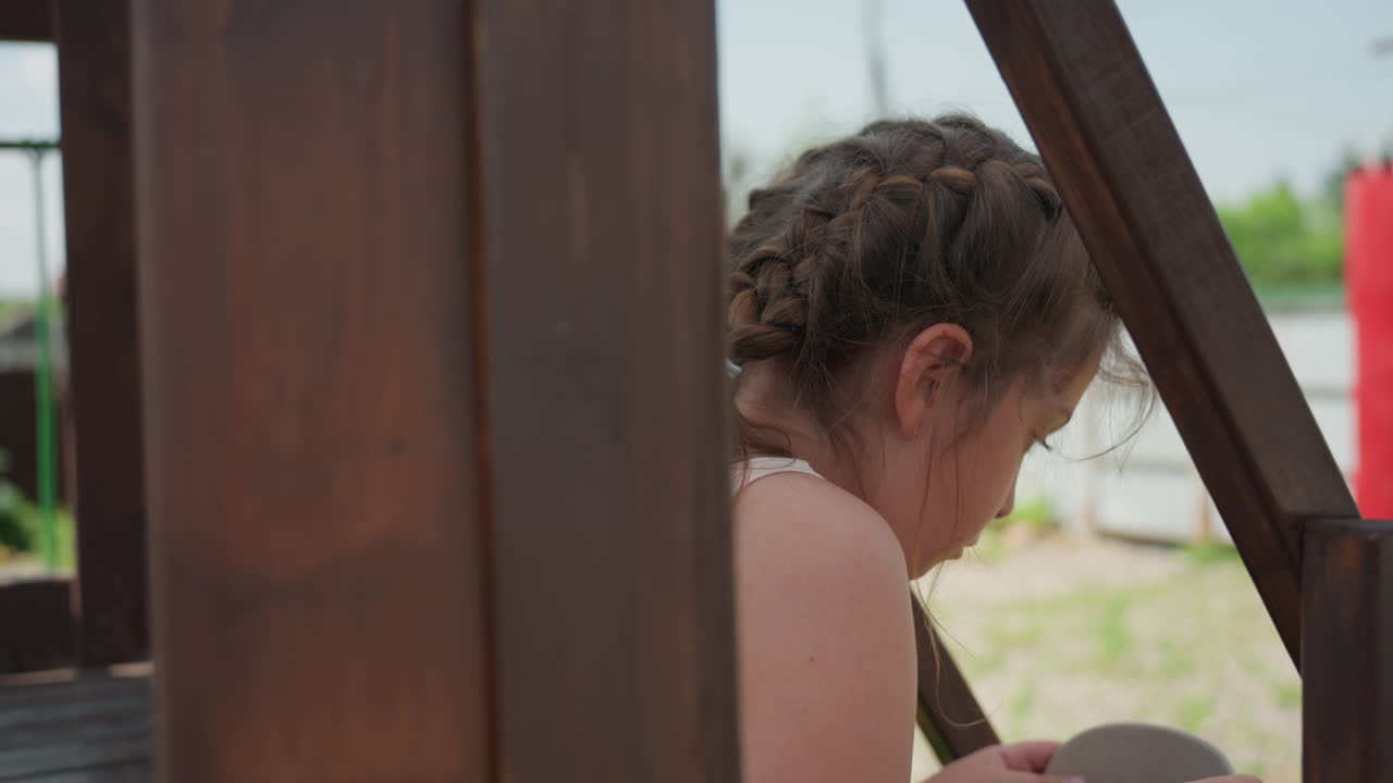 Child Sitting Behind Wooden Railing, Braided Hair In Profile, Summer Backyard Porch Scene With Soft Sunlight And Gentle Shade, Contemplative Mood As Child Studies Hands And Surroundings, Intimate
