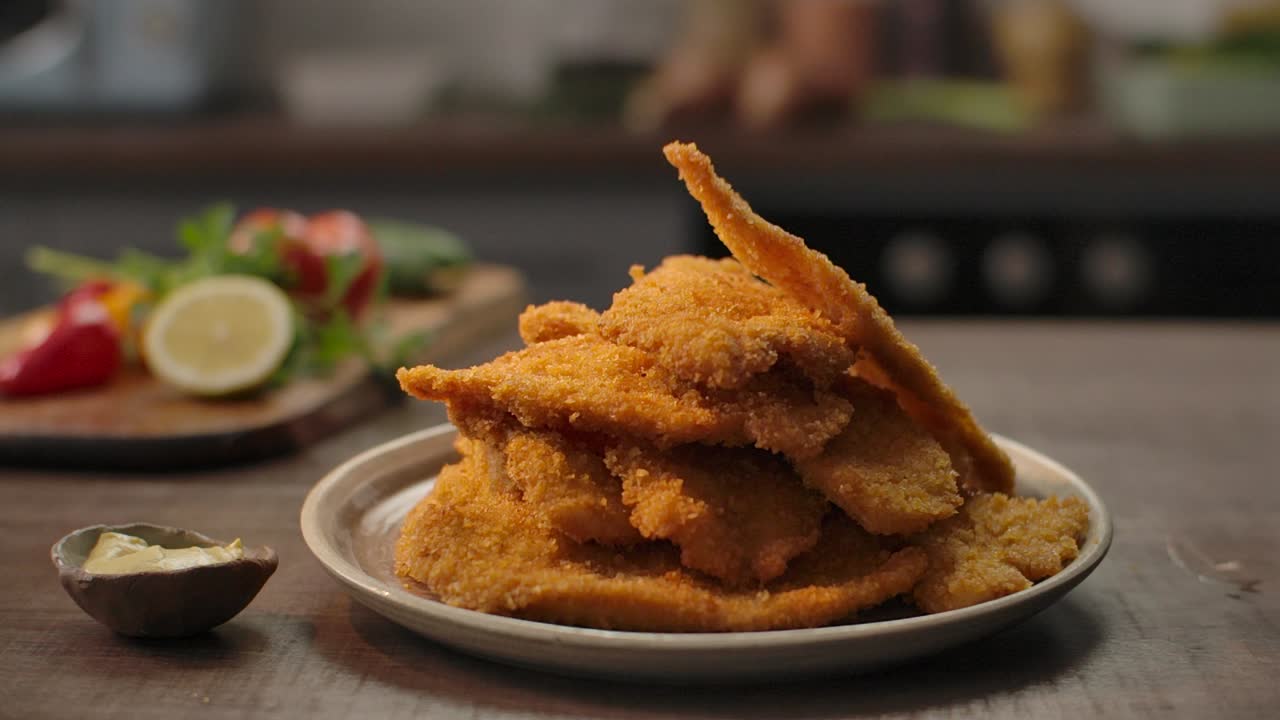Close up of a chef stacking perfectly breaded, golden-brown chicken schnitzels on a ceramic plate, styled with fresh lemon and peppers in a cozy kitchen