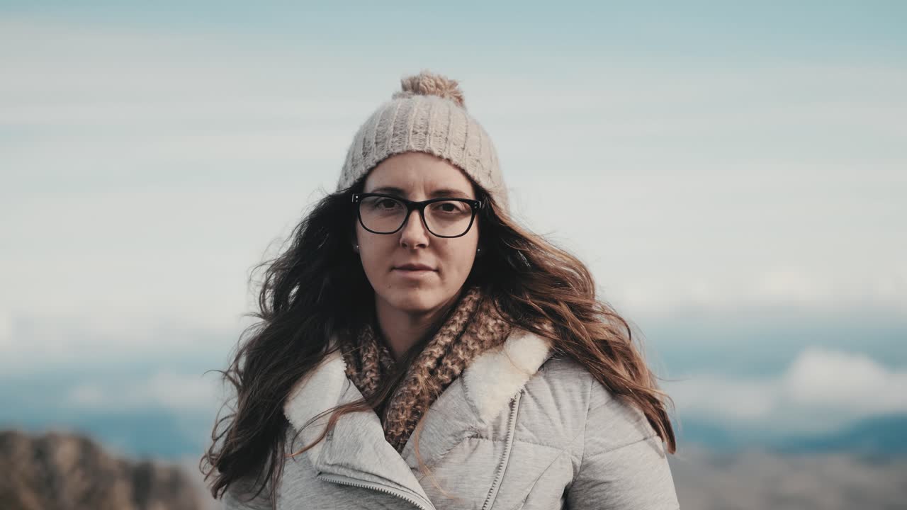 Nahaufnahme einer jungen Frau mit Brille und langen braunen Haaren, nachdenklich in die Kamera blickend, vor einer atemberaubenden Berg- und Wolkenhimmelkulisse