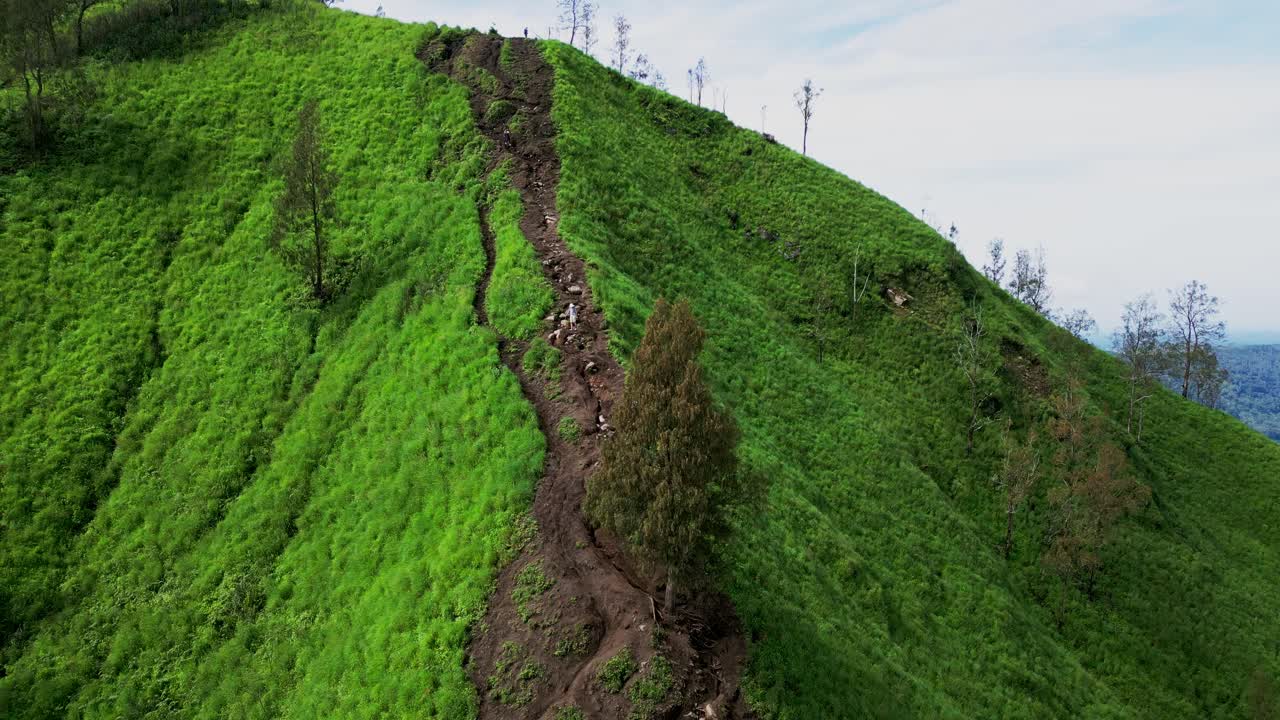 In the golden light of early morning, hikers descend Trunyan Hill’s vibrant green landscape, filmed from above by drone, revealing a serene and untouched tropical countryside in Bali.Mountain