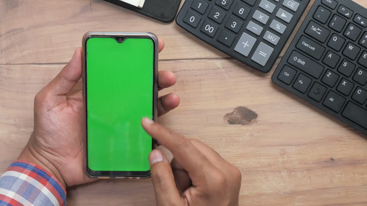 Person holding a smartphone with a green screen, on a wooden desk with a calculator and keyboard