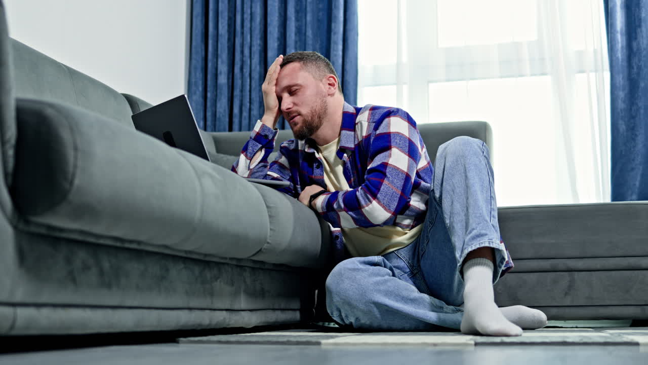 Uomo seduto per terra vicino a un divano, che usa un laptop in salotto di giorno, indossando abiti casual e pensando a qualcosa di importante per lui.