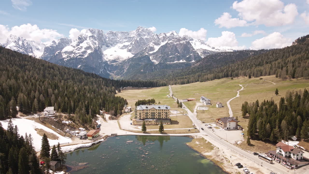 Camera descends to reveal a grand lakeside hotel framed by evergreen forest, thawing lake, and snow‑capped Dolomite peaks under a bright spring sky