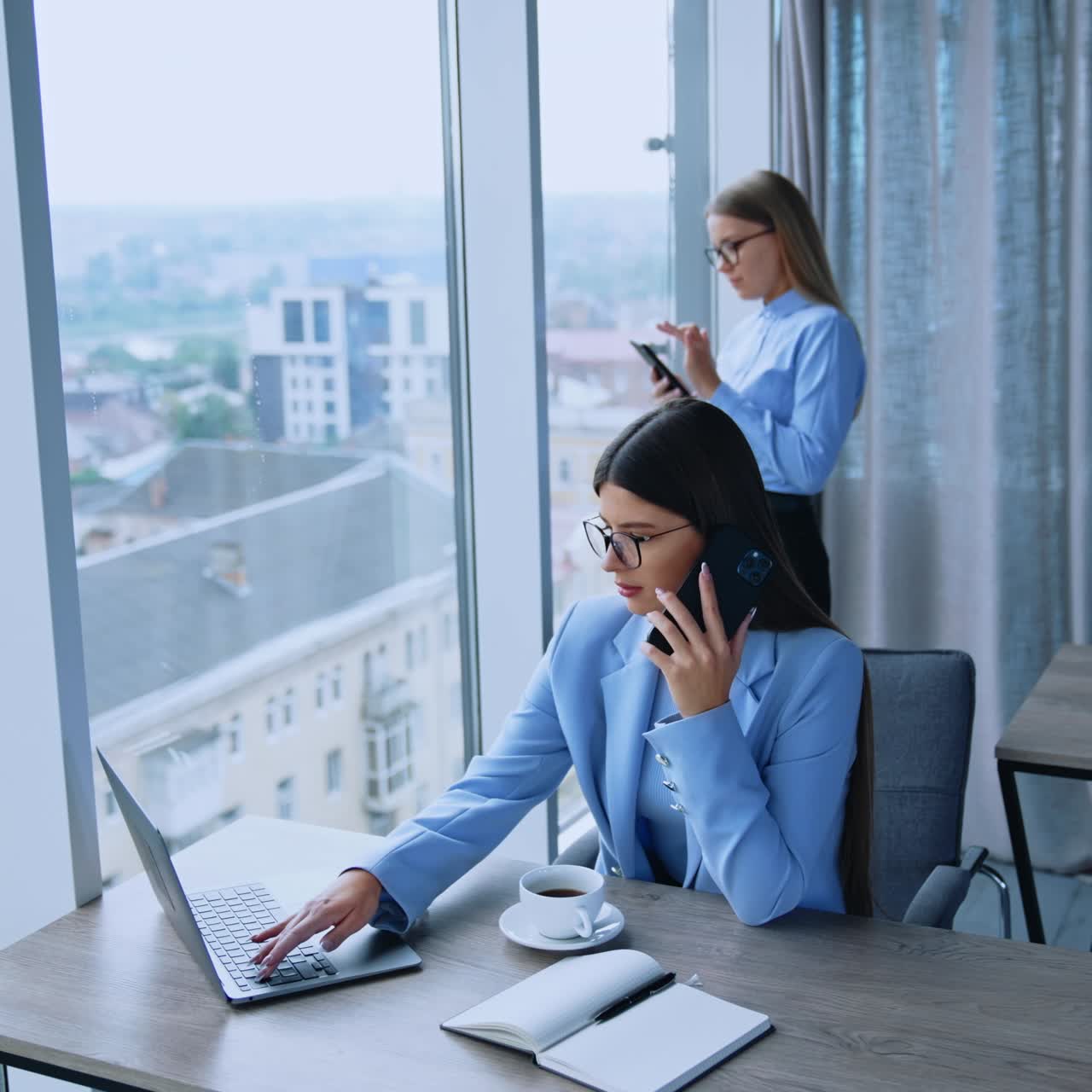 Office employees having productive day at work. Female colleagues using phones and laptop. Office with panoramic windows at backdrop