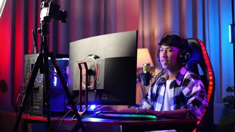 asian boy streamer smiling and crossing his arms while playing game over network on personal computer. live stream video game, desk illuminated by rgb led strip light