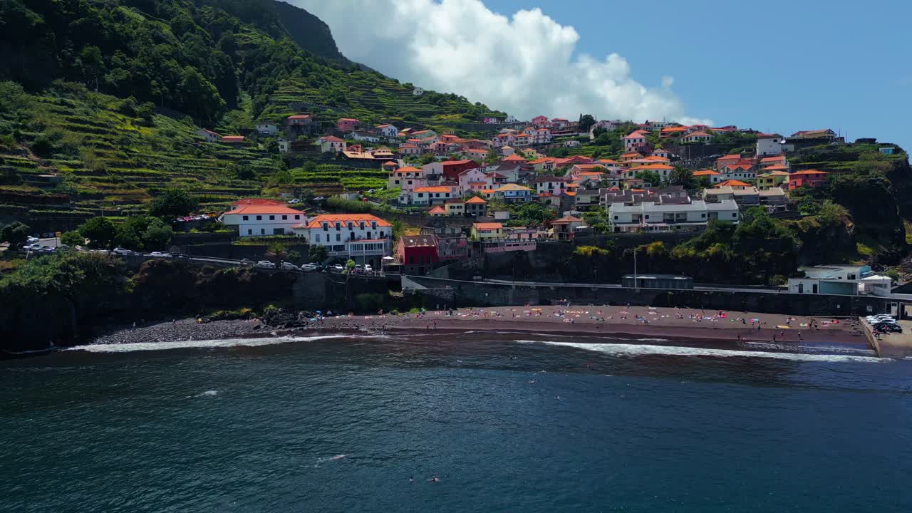 Vista aérea costeira da vila litorânea de Seixal, na Madeira, Portugal, mostrando casas nas encostas, paisagens em socalcos, pessoas na praia de areia e águas calmas do Oceano Atlântico