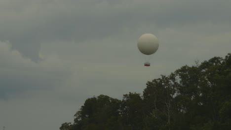 a large white hot air balloon with a red gondola floats above the treetops near niagara falls under a cloudy sky, offering a peaceful aerial perspective