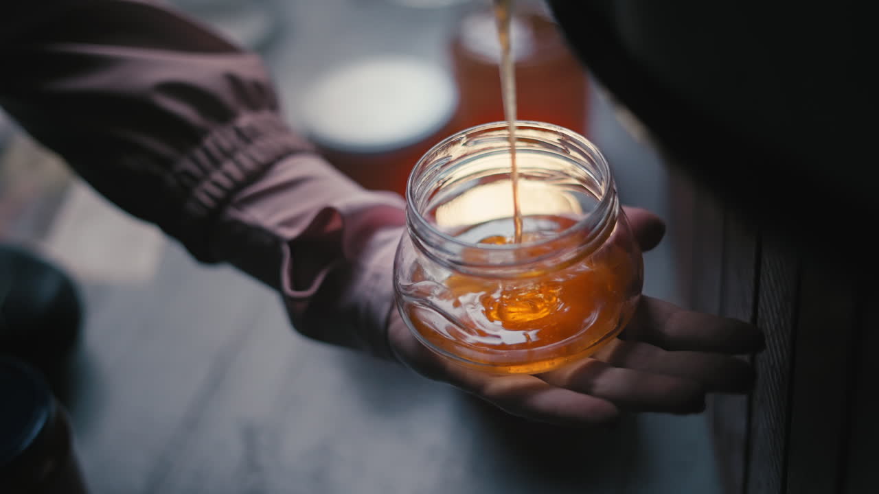 Mano che tiene un barattolo di vetro, versando e riempiendo miele biologico liquido estratto fresco e dorato, produzione tradizionale di miele, apicoltura e allevamento di api in un apiario