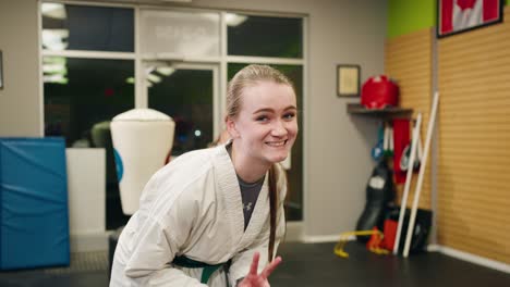 A-Tired-Young-Woman-Teenage-Girl-Wearing-Green-Belt-has-Fun-Smiling-Laughing-Peace-Sign-While-Taekwondo-Martial-Arts-Training-Studio-Inside-a-Gym-Slowmo-Black-Belt-Master-Background-Punching-Bag