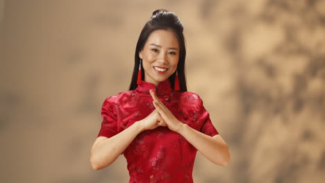 Portrait-of-young-Asian-woman-in-red-traditional-clothes-smiling-cheerfully-at-camera-and-doing-greeting-gesture-with-hands