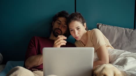 the guy and the girl are looking at the laptop screen while lying on the bed against the background of a turquoise wall in the morning. the couple jointly decides on the purchase of goods in the online store