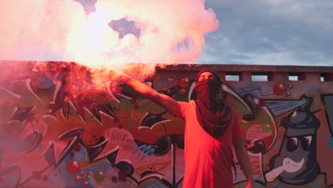 young man in balaclava with red burning signal flare on the roof with graffiti background, slow motion