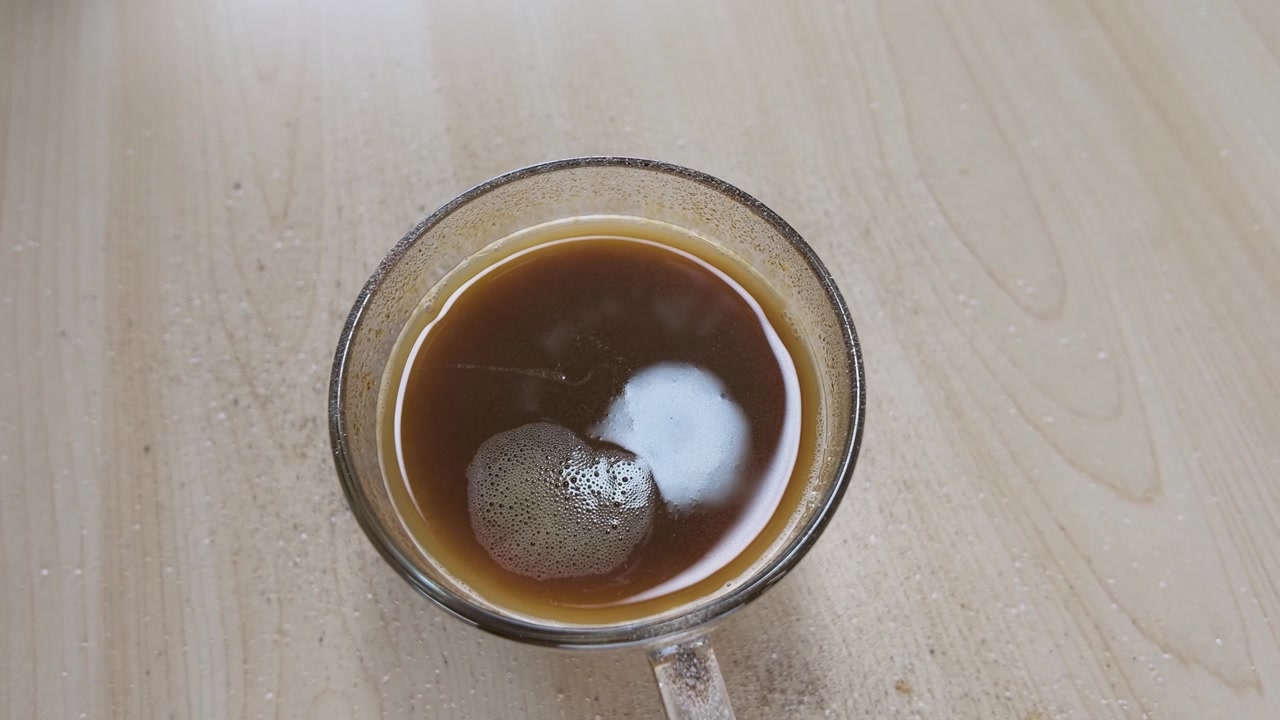 Zucchero bianco granulato cade in una tazza di vetro di caffè nero su un tavolo di legno