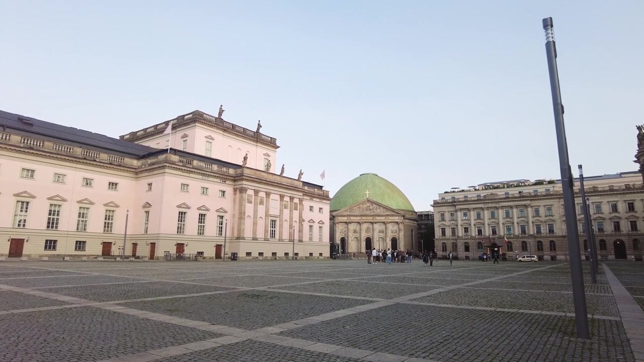 Uma vista panorâmica da praça de paralelepípedos da histórica Bebelplatz revela uma ópera neoclássica e uma catedral com cúpula, enquanto pedestres caminham ao longe