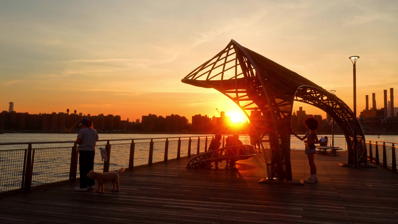 New York, USA, 1. juli 2026: Buet ståltag ved East River ved solnedgang. Bagbelyste besøgende samles på en træmole, mens orange sollys indrammer havnefrontens struktur.