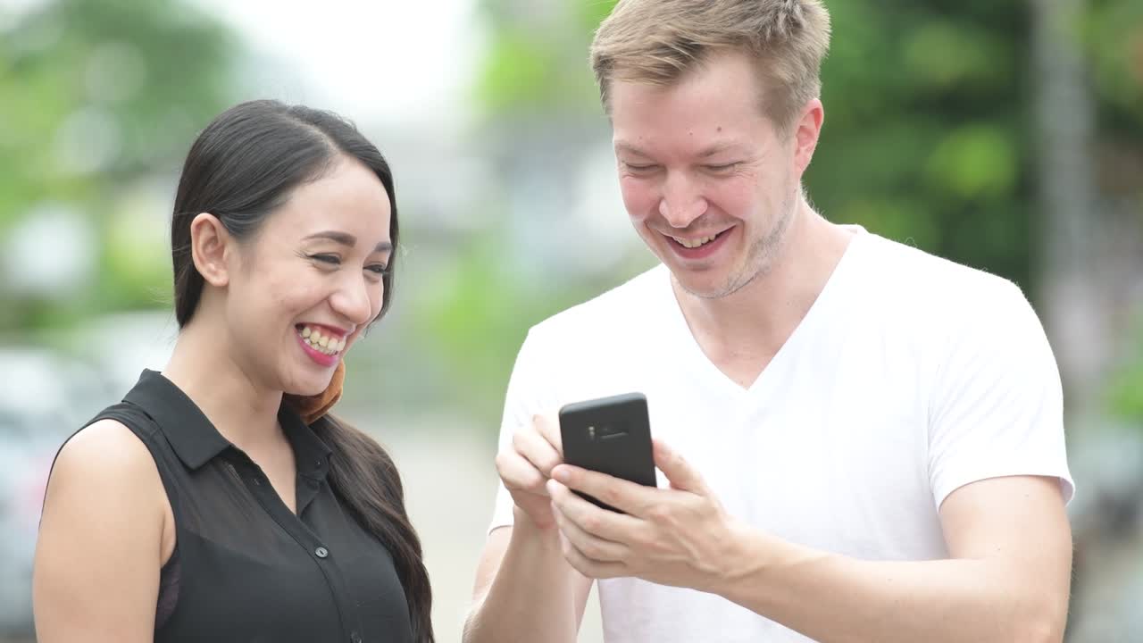 젊은 다민족 부부가 거리에서 함께 전화를 사용하고 있습니다.