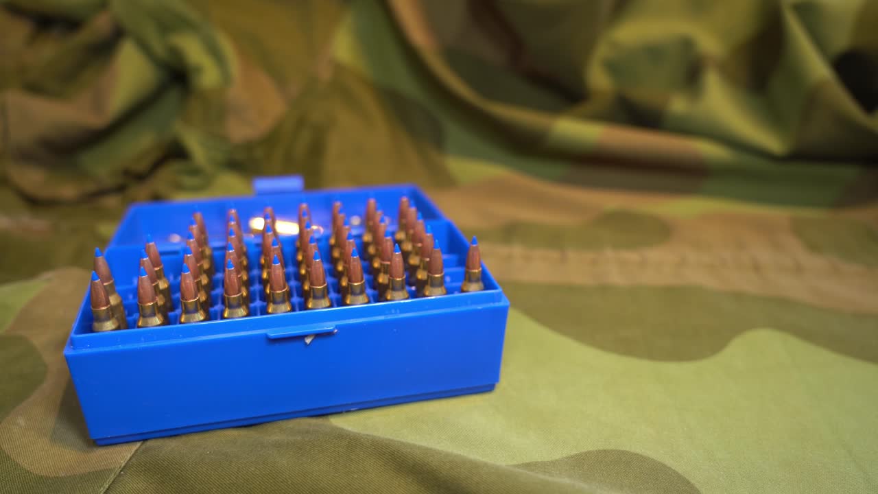 Ammunition box full of powerful military grade 223 caliber varmint bullets with blue plastic tip laying on camouflage jacket - Slider from left to right