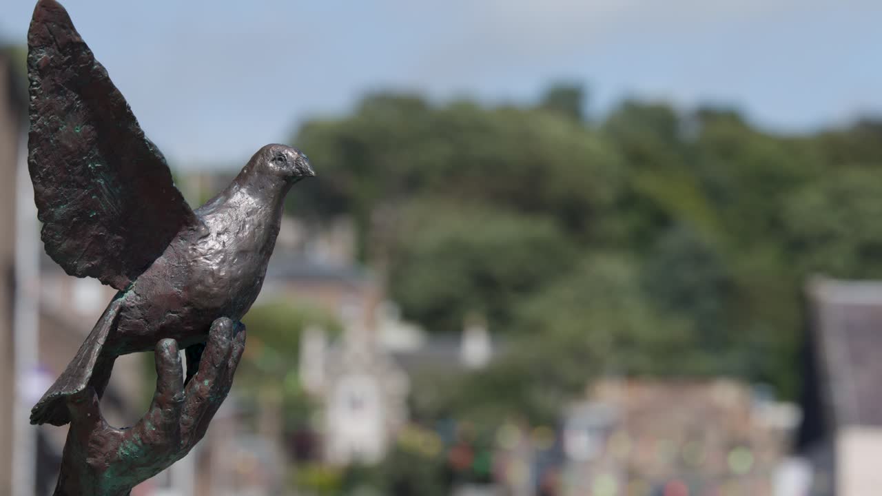 Static close-up of bronze dove sculpture with blurred village and greenery in bright daylight