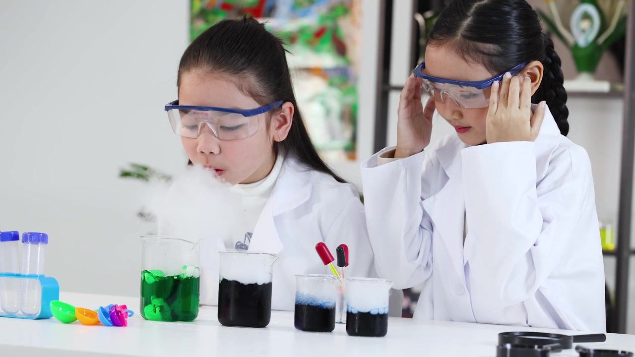 Two young girls wear safety goggles while observing chemical reactions and smoke in beakers