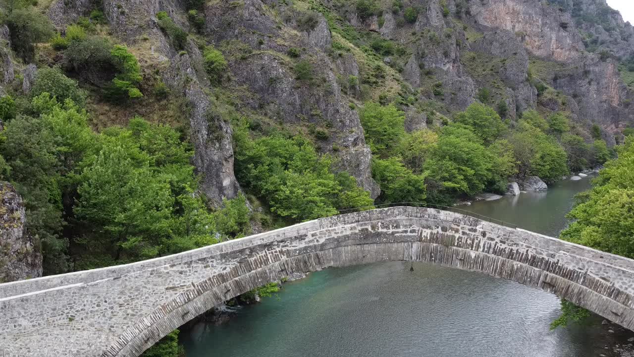 Cinematic drone descent over the historic Konitsa stone bridge. A stunning bird's-eye view of the largest single-arch bridge in the Balkans, set at the entrance of the Aoos Gorge, Epirus