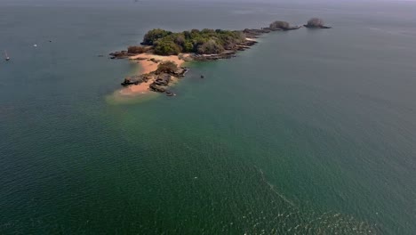 Vuelo-Hacia-Atrás-De-La-Pintoresca-Isla-Con-Cala-De-Playa-Rocosa-Y-Veleros-Cercanos