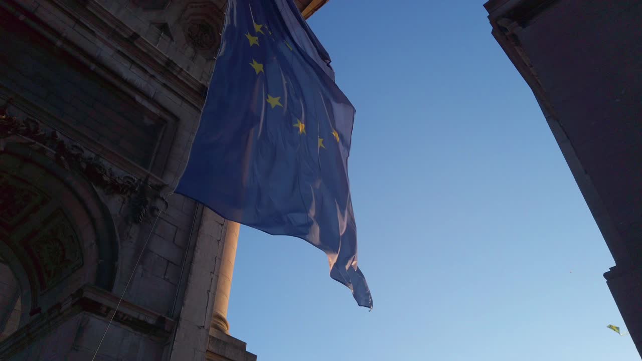 Euroopan lippu liehumassa kivirakennusten välissä matalasta kuvakulmasta auringonlaskun aikaan.