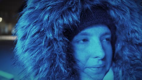 mujer joven con una capucha de piel cubierta de nieve mirando a su alrededor frente a una luz azul en un paisaje invernal por la noche - primer plano