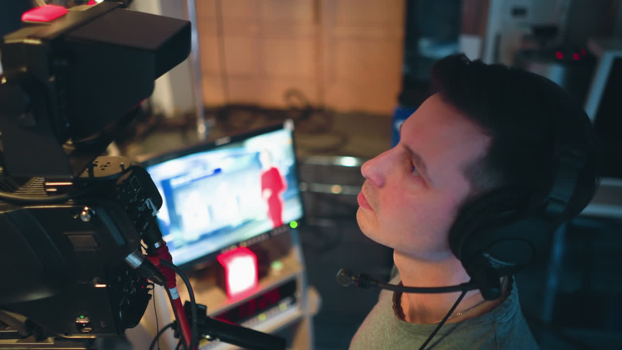 Male cameraman wearing headset concentrates while operating television camera in control room during live studio broadcast, with presenter in red dress visible on small monitor and multiple production screens in background