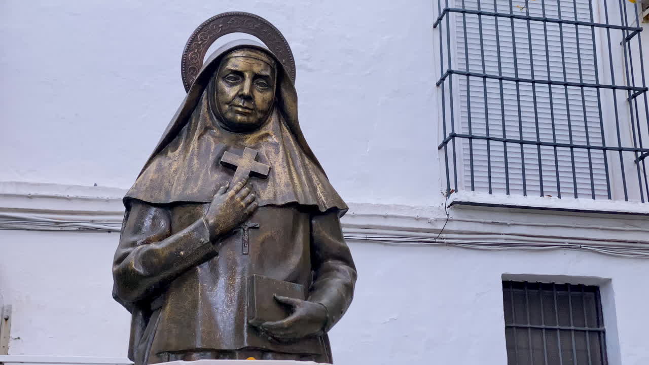 Escultura de bronze de uma freira religiosa segurando uma cruz e um livro em Sanlúcar de Barrameda, Espanha, contra a parede de um edifício branco.