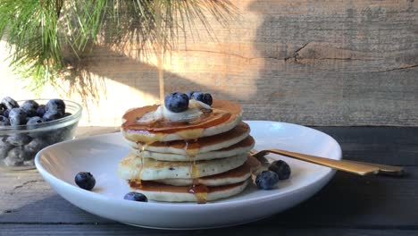 Side-view-of-stack-of-blueberry-pancakes-with-pouring-syrup-drizzled-over-sides-in-morning-sunlight