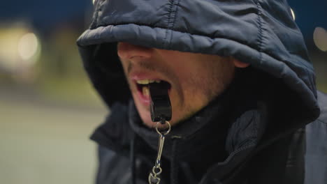 close-up of a man holding a whistle in his mouth, partially obscured by a hooded jacket, the background is blurred and dimly lit