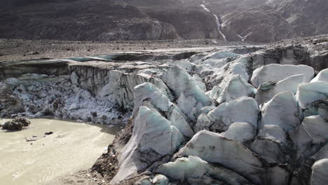 Panoramic-view-of-melting-glacier-covered-in-moraine,-Muddy-glacier-lake-beneath-mountains-in-summer,-Aerial-closeup