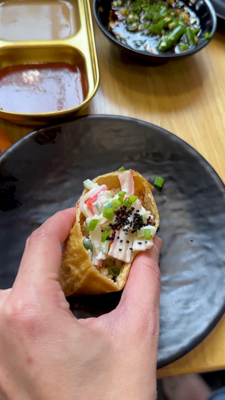 Hand grabs seafood inari sushi roll from black plate, natural lighting, handheld camera, restaurant setting