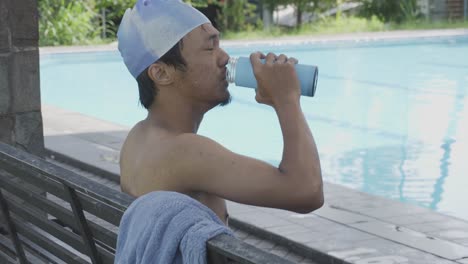 young asian male athlete sitting by the pool, drinking, wipe himself with towel and resting after swimming