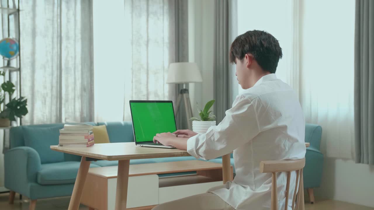 Side View Of Asian Man Student Typing On Green Screen Computer At Home