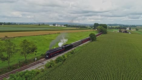 An-Aerial-View-Traveling-In-Front-of-An-Antique-Steam-Passenger-Train-Blowing-Smoke-Traveling-Across-a-Crossing-and-Thru-Rich-Farmlands-on-a-Summer-Day