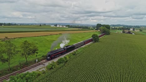 an aerial view traveling in front of an antique steam passenger train blowing smoke traveling across a crossing and thru rich farmlands on a summer day