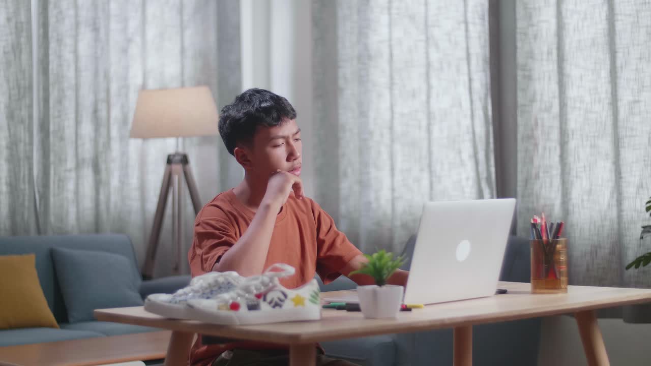 아시아 소년 신발 디자이너가 집에서 노트북으로 일하면서 생각하고 있습니다.