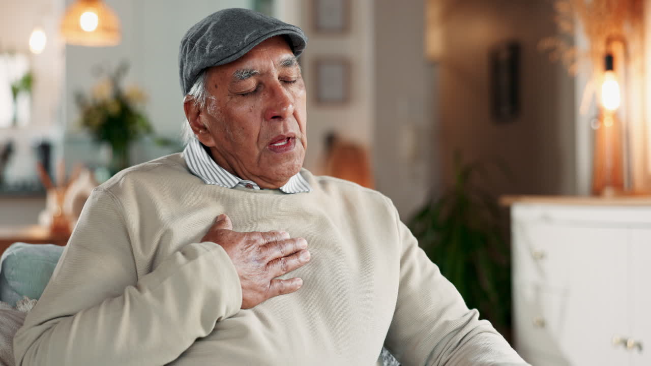 A senior man sitting on a couch in a living room, looking down and holding his chest, showing signs of being sick and uncomfortable.