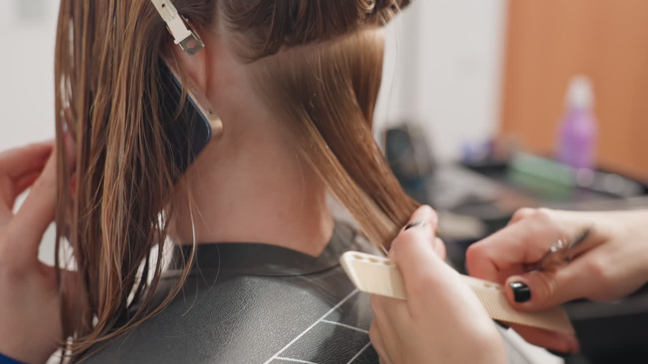 Primer plano de un corte de pelo de precisión en la nuca a un cliente caucásico con el estilista separando mechones, clips y peine en el encuadre, primer plano de las manos usando tijeras y cizallas, capa de peluquería y estación de trabajo visibles, técnica centrada